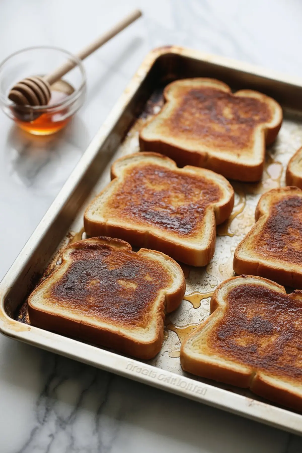 Tray of baked cinnamon toast slices with caramelized cinnamon sugar tops, surrounded by drizzles of melted butter and a small glass bowl of honey with a wooden honey dipper.
