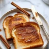 Two slices of cinnamon toast drizzled with honey on a round plate, surrounded by cinnamon sticks and sprinkled cinnamon powder, styled with a vintage butter knife on a marble surface.