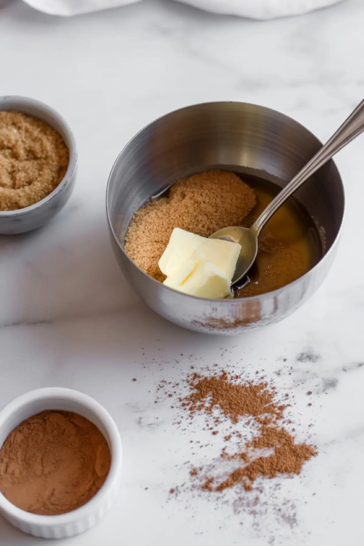 Mixing bowl with softened butter, brown sugar, cinnamon, and vanilla extract for making the cinnamon filling, surrounded by scattered spices on a white marble counter.