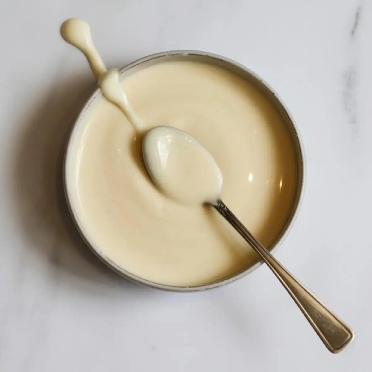Bowl of creamy vanilla glaze with a spoon, used for drizzling over puff pastry cinnamon twists, placed on a clean white surface for a minimal baking aesthetic.