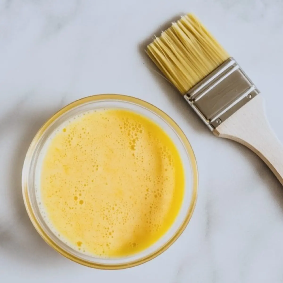 Small bowl of beaten egg yolk next to a pastry brush, ready for egg wash application on puff pastry before baking cinnamon twists for a shiny, golden finish.