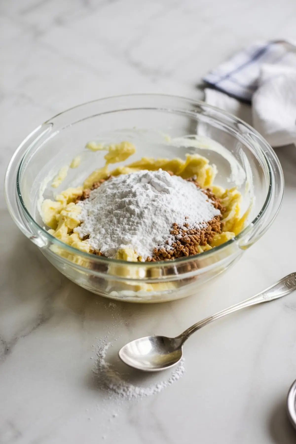 Glass mixing bowl with softened butter, brown sugar, and powdered sugar on a marble countertop, ready to be creamed for cookie dough frosting preparation.
