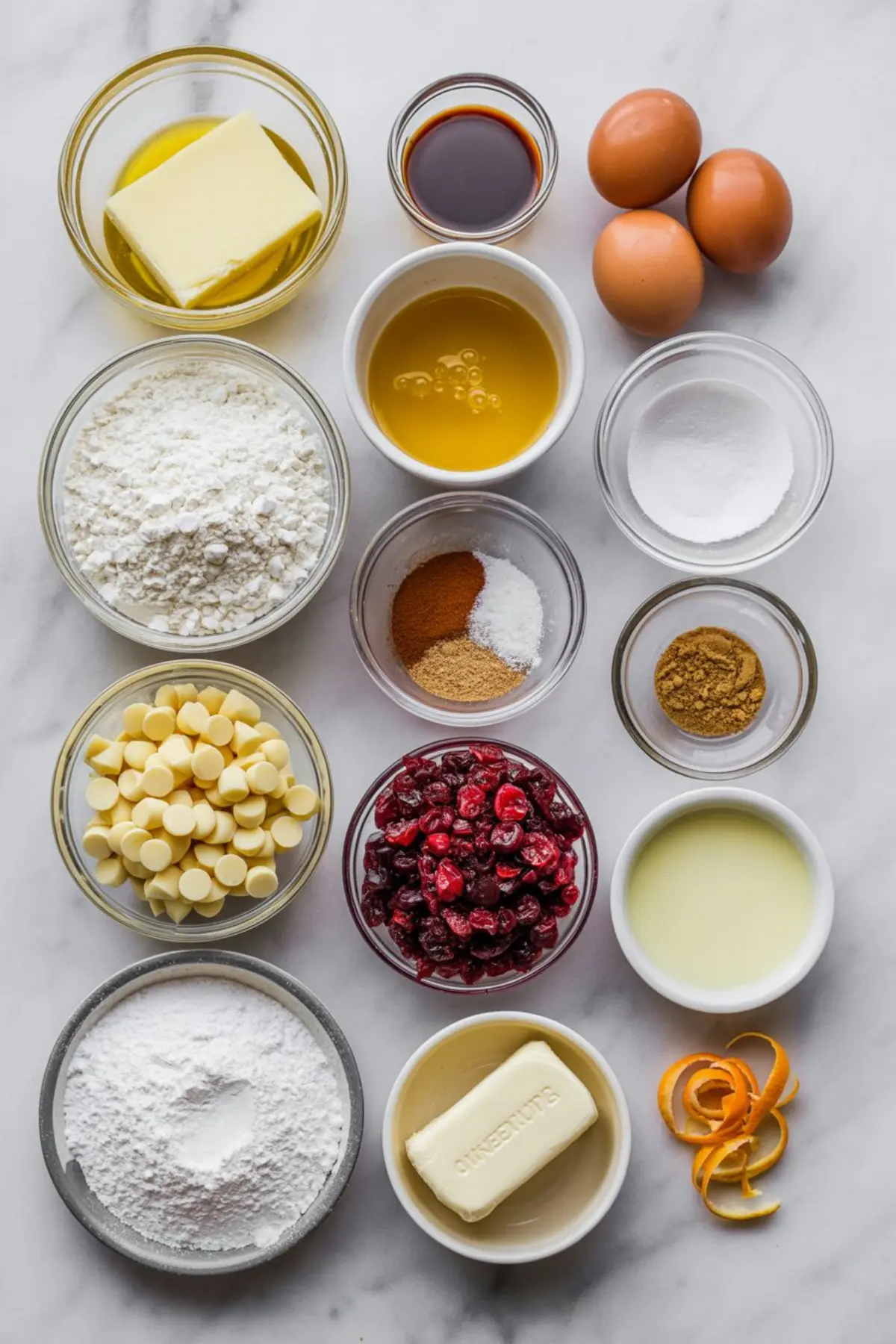 Flat lay of baking ingredients in glass bowls including butter, flour, white chocolate chips, dried cranberries, spices, eggs, powdered sugar, and orange zest, arranged on a marble background.
