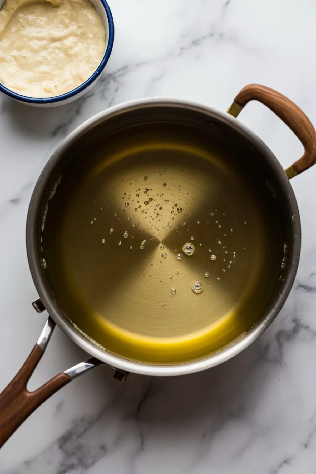 A large stainless steel pot filled with oil heating on the stove, prepared for frying apple fritters, with a bowl of batter beside it.
