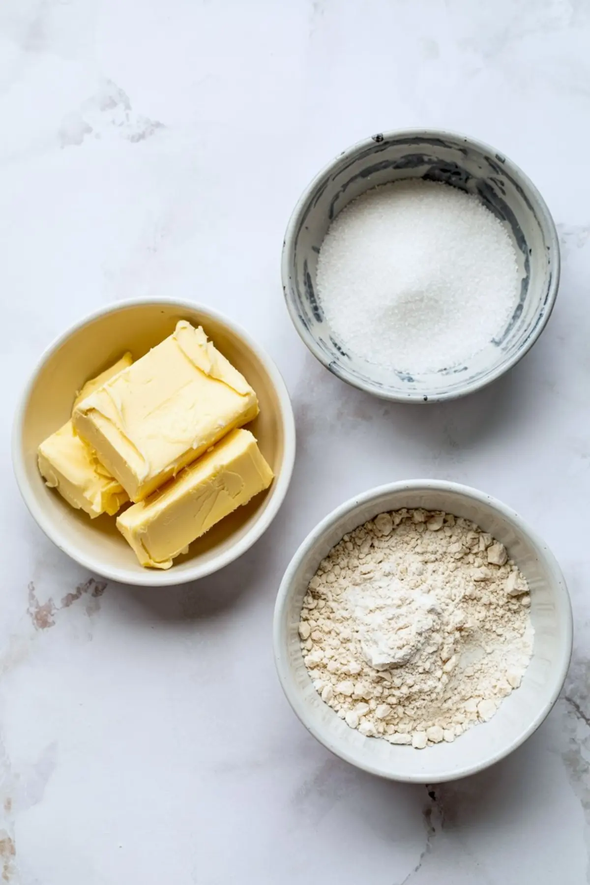 Top view of three baking ingredients in ceramic bowls: softened butter, white granulated sugar, and all-purpose flour on a marble surface.