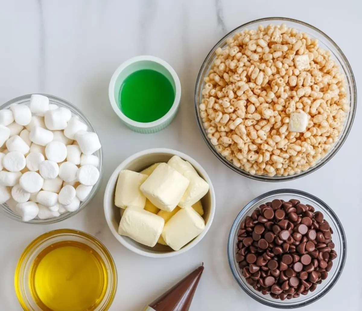 Flat lay of Halloween Rice Krispie treat ingredients including mini marshmallows, green food coloring, rice cereal, chocolate chips, butter, vanilla extract, and a piping bag with chocolate icing.
