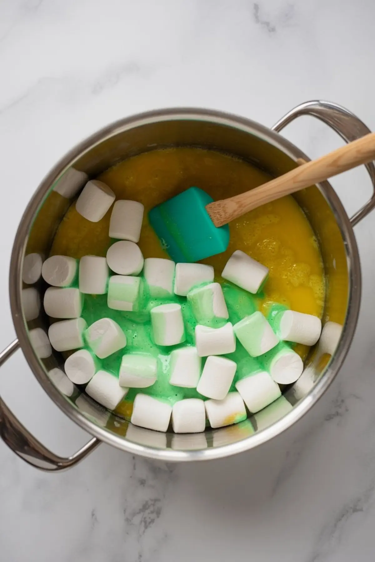 Stainless steel pot filled with melted butter, green food coloring, and whole mini marshmallows being stirred with a turquoise spatula, preparing the base for Rice Krispie treats.
