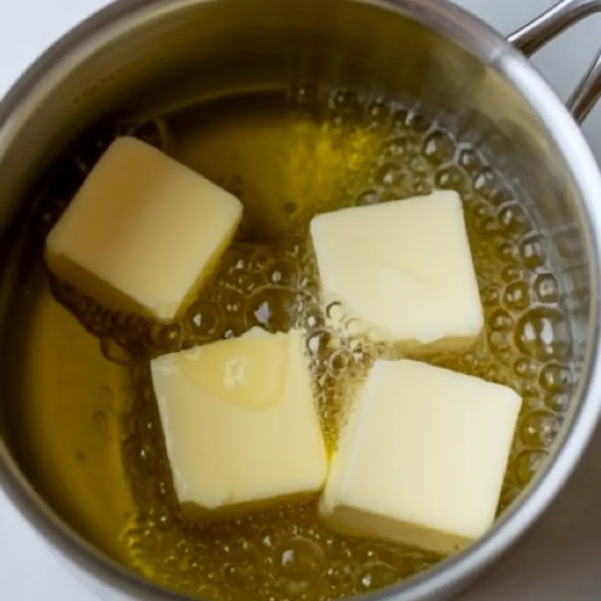 Close-up of butter cubes melting in a saucepan, with bubbling edges indicating the start of a cooking process on a smooth surface background.