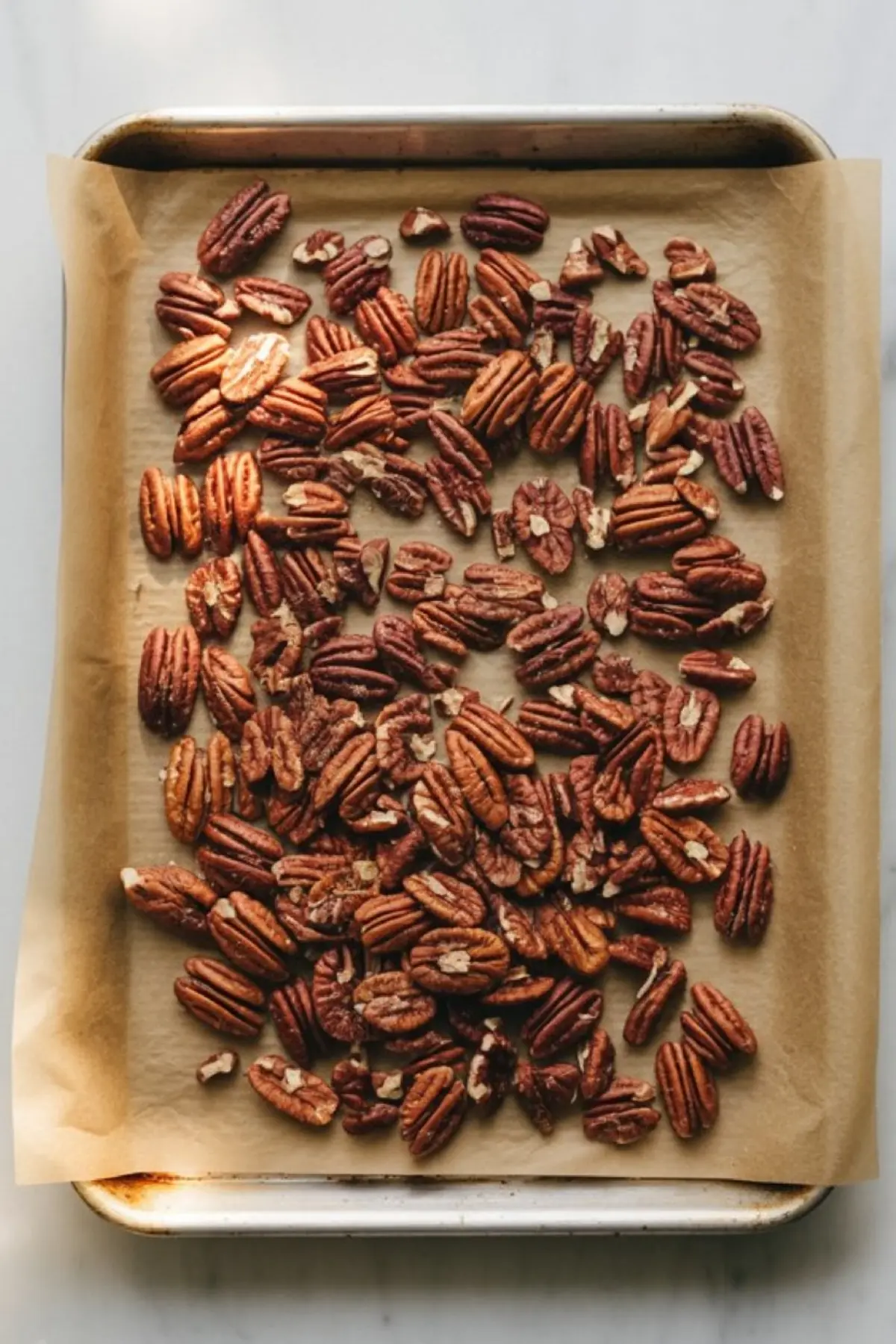 Baking sheet lined with parchment paper topped with a single layer of raw pecan halves, prepared for roasting on a white marble background.