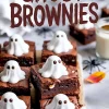 Ghost-themed Halloween brownies on a tray, each square topped with a piped white ghost made of meringue and chocolate chip eyes, styled with black spider decorations and a glass of milk.