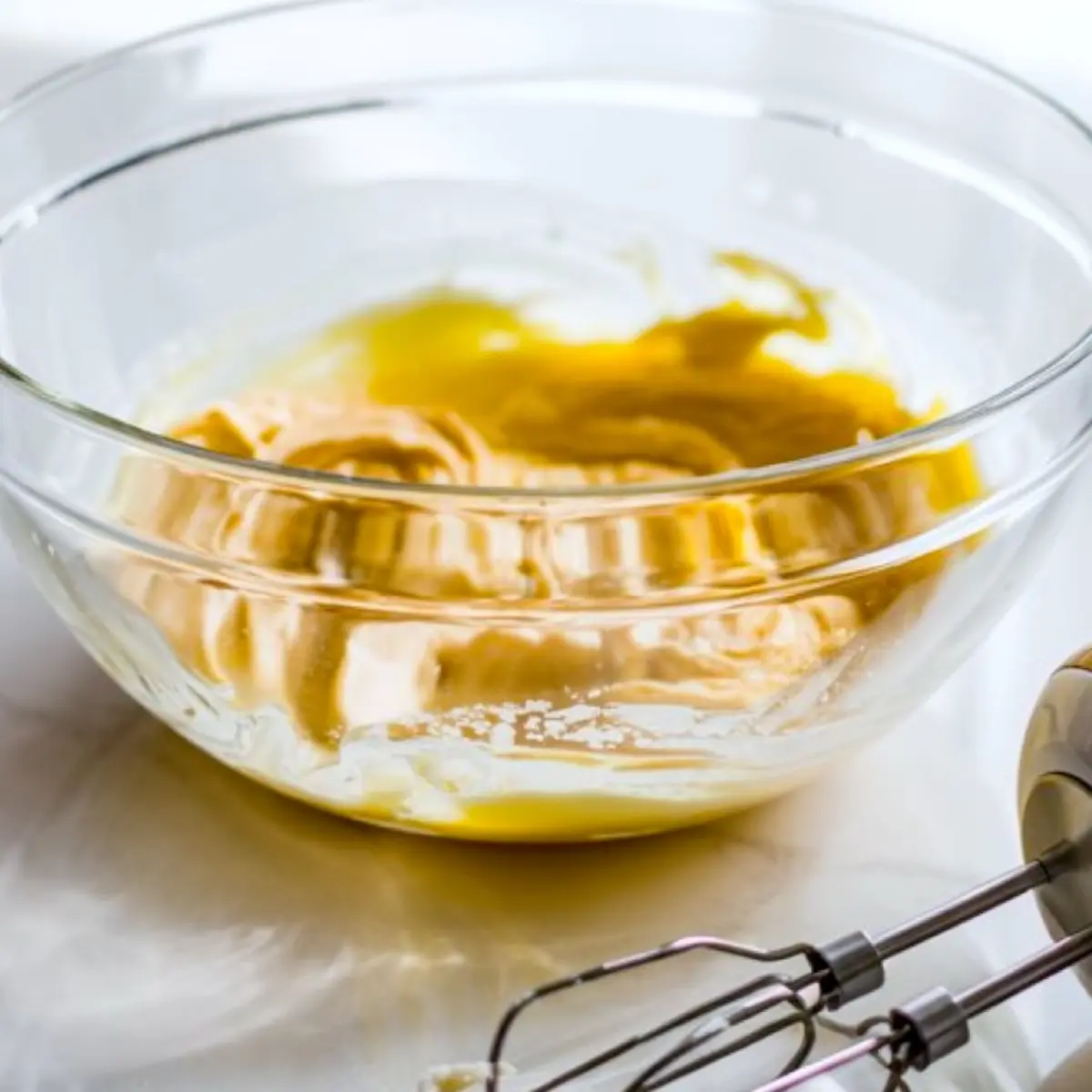 Creamy yellow egg and sugar mixture partially whipped in a glass bowl with electric mixer attachments resting beside it on a marble counter.
