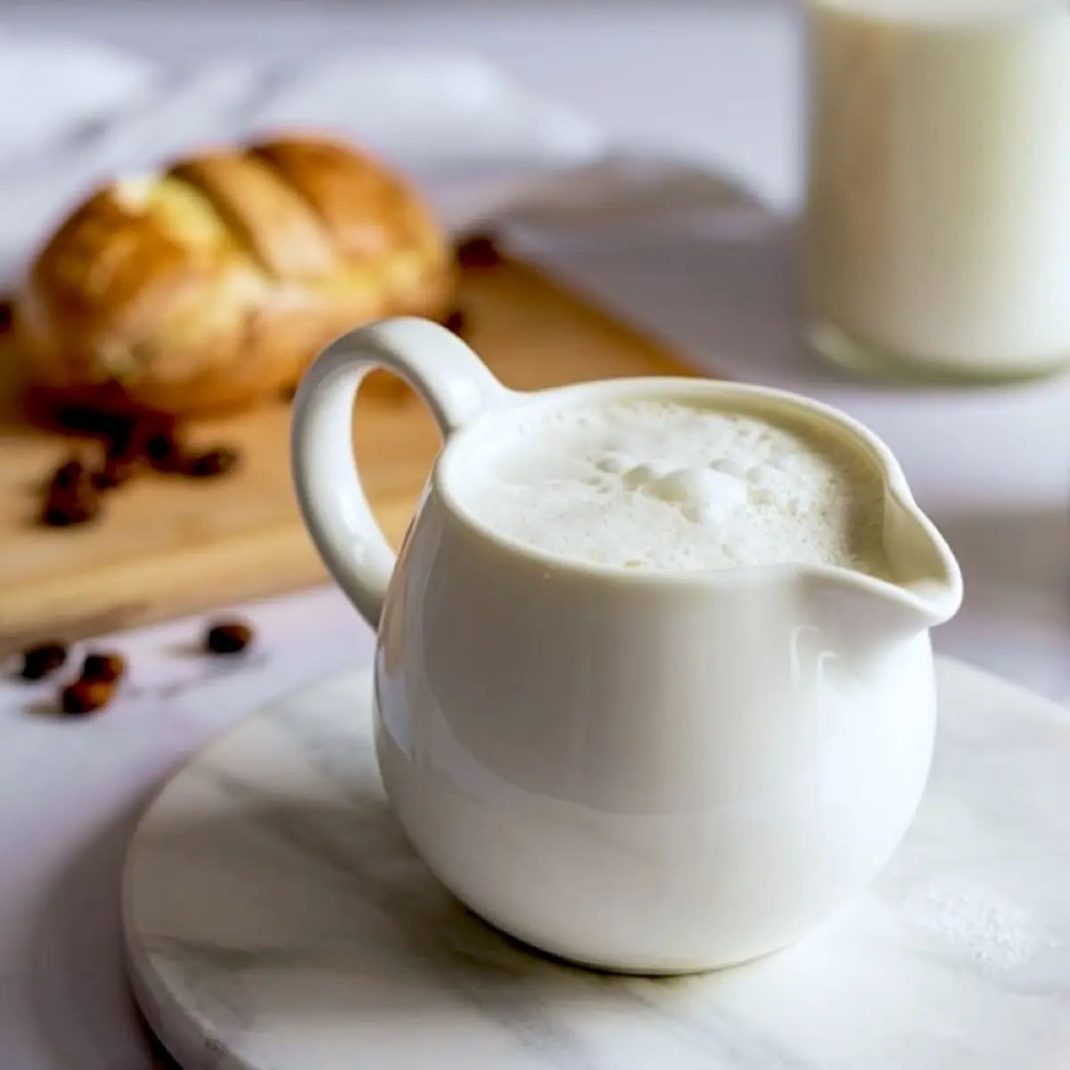 White ceramic milk frothing pitcher filled with steamed milk foam, placed on a marble board with bread and coffee beans in the background.