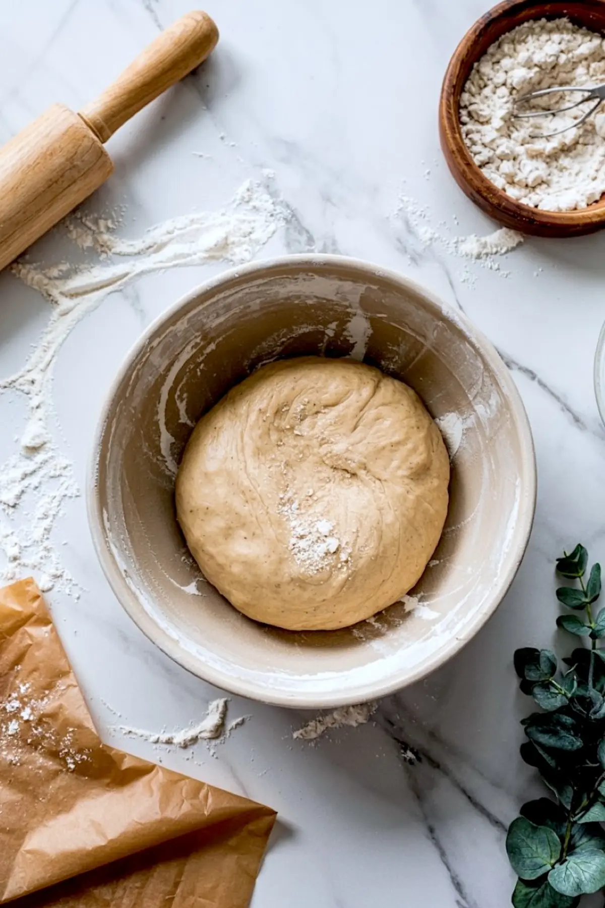 Round ball of risen dough resting in a flour-dusted ceramic bowl on a marble countertop, surrounded by a wooden rolling pin, bowl of flour, brown parchment paper, and fresh eucalyptus.
