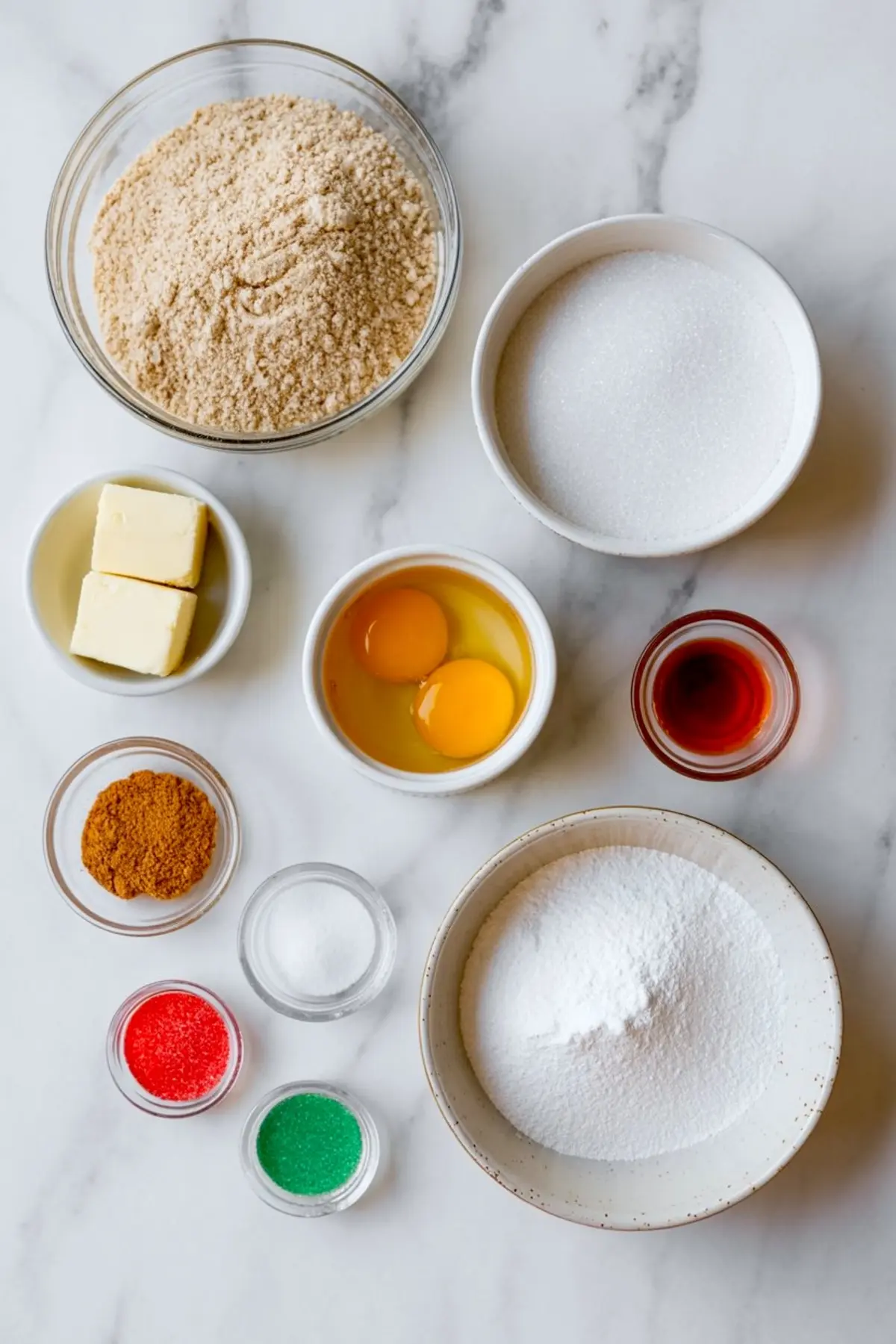 Flat lay of snickerdoodle cookie ingredients on a marble surface, including almond flour, granulated sugar, butter, eggs, vanilla extract, cinnamon, baking soda, and red and green sanding sugars.

