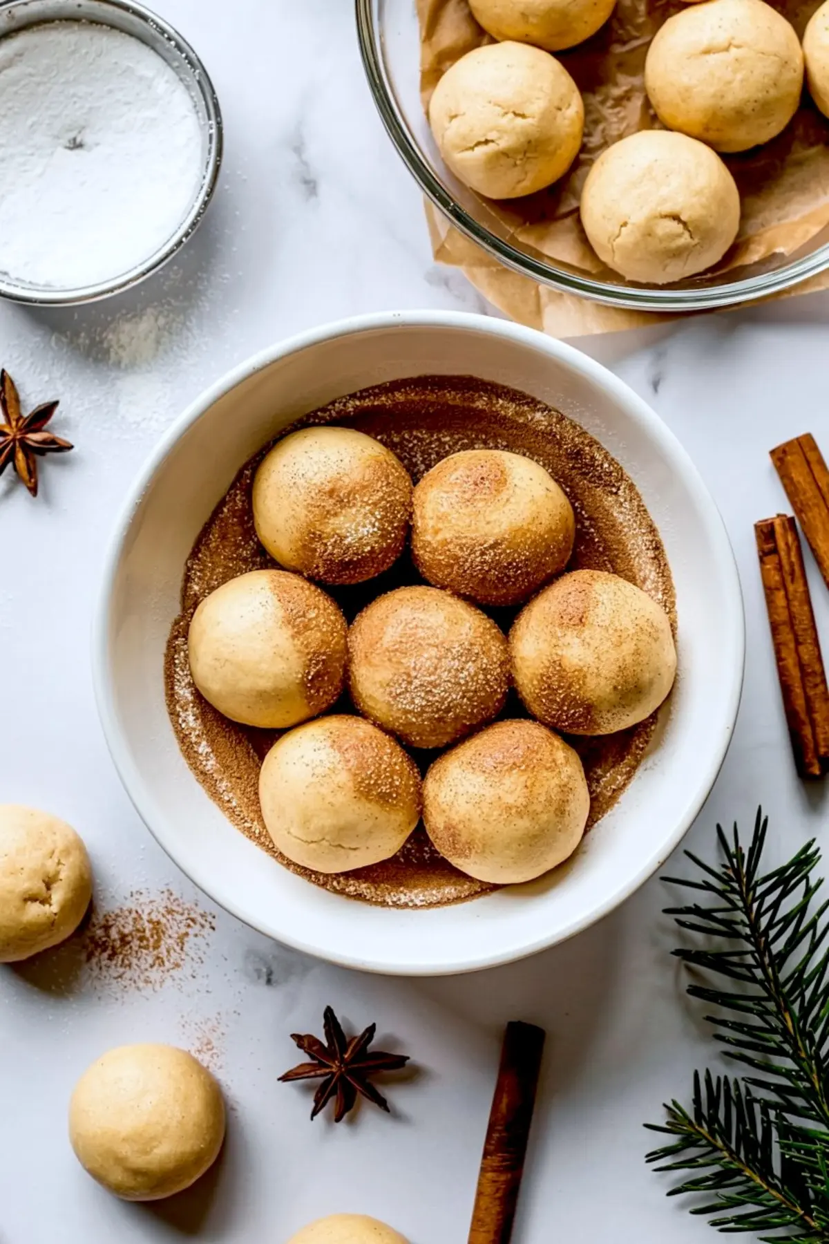 Round cookie dough balls rolled in cinnamon sugar arranged in a bowl, surrounded by spices and evergreen sprigs on a white background for holiday baking prep.
