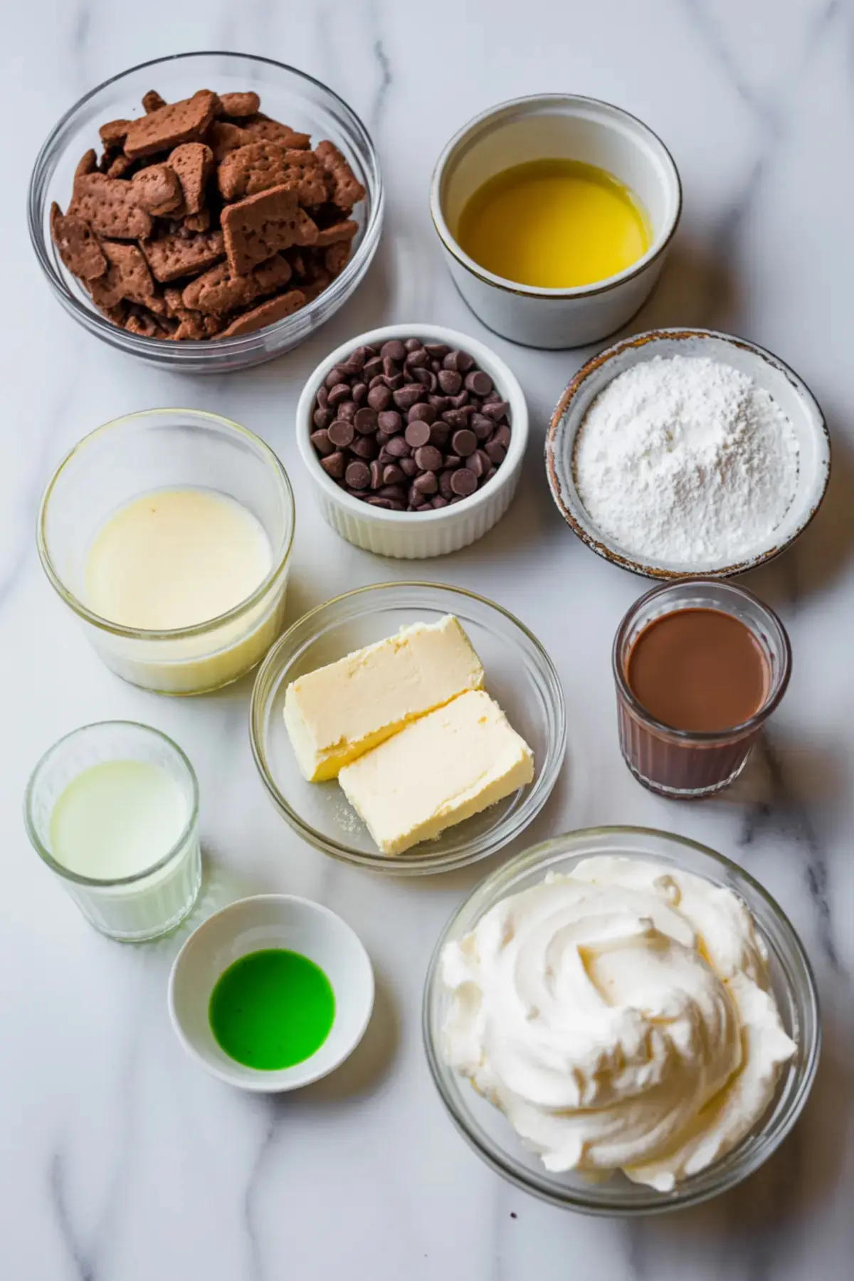 Flat lay of grasshopper pie ingredients including chocolate graham crackers, chocolate chips, powdered sugar, melted butter, heavy cream, mint extract, whipped topping, sweetened condensed milk, and softened butter arranged in clear bowls on a marble countertop.