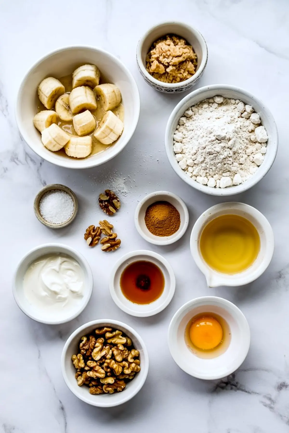 Flat lay of banana walnut muffin ingredients on a marble surface, including chopped bananas, flour, brown sugar, Greek yogurt, walnuts, peanut butter, cinnamon, salt, vanilla extract, egg, and vegetable oil in individual white bowls.