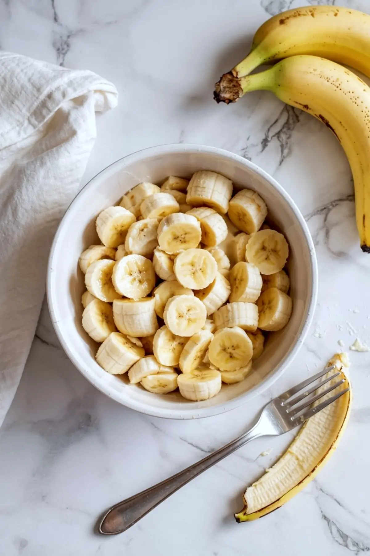 White ceramic bowl filled with freshly sliced ripe bananas on a marble surface, with a silver fork and banana peel nearby, and whole bananas resting beside a white kitchen towel.