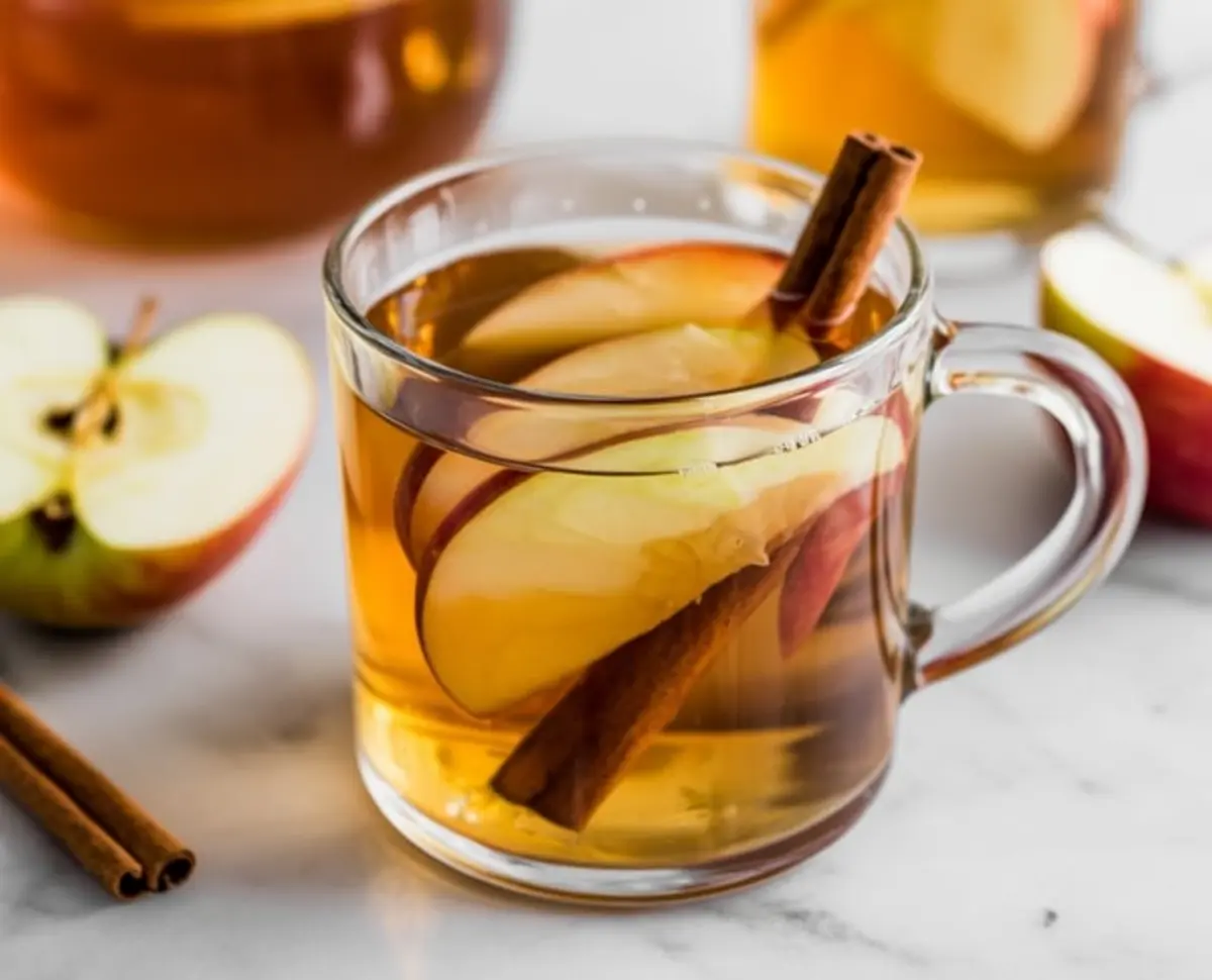 Glass mug of hot apple cider garnished with fresh apple slices and a cinnamon stick, placed on a marble surface next to halved apples and loose cinnamon sticks.