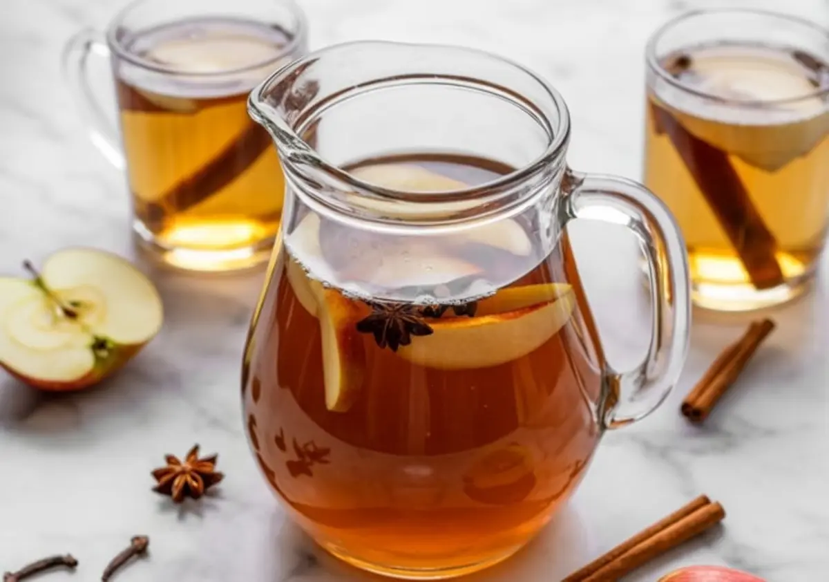 Glass pitcher of apple cider infused with apple slices and star anise, surrounded by mugs filled with cider, cinnamon sticks, and halved apples on a white marble countertop.