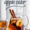 Pitcher of apple cider filled with cinnamon sticks and sliced red apples, surrounded by glasses of cider and the text “homemade apple cider” over a white curtain background.