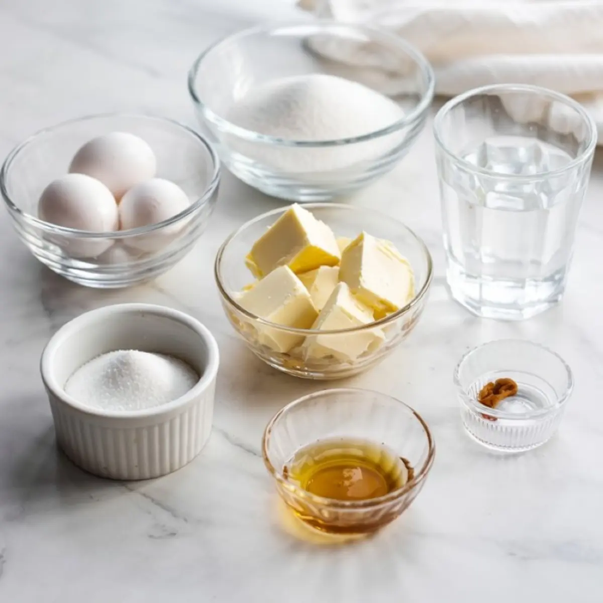 Flat lay of Italian buttercream ingredients arranged on a marble countertop, including whole eggs, granulated sugar, unsalted butter cubes, water, vanilla extract, and cream of tartar in small glass bowls.