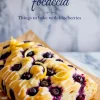 Three-quarter view of baked lemon blueberry focaccia loaf with glossy blueberries, white glaze, and curled lemon zest on parchment-lined marble surface.