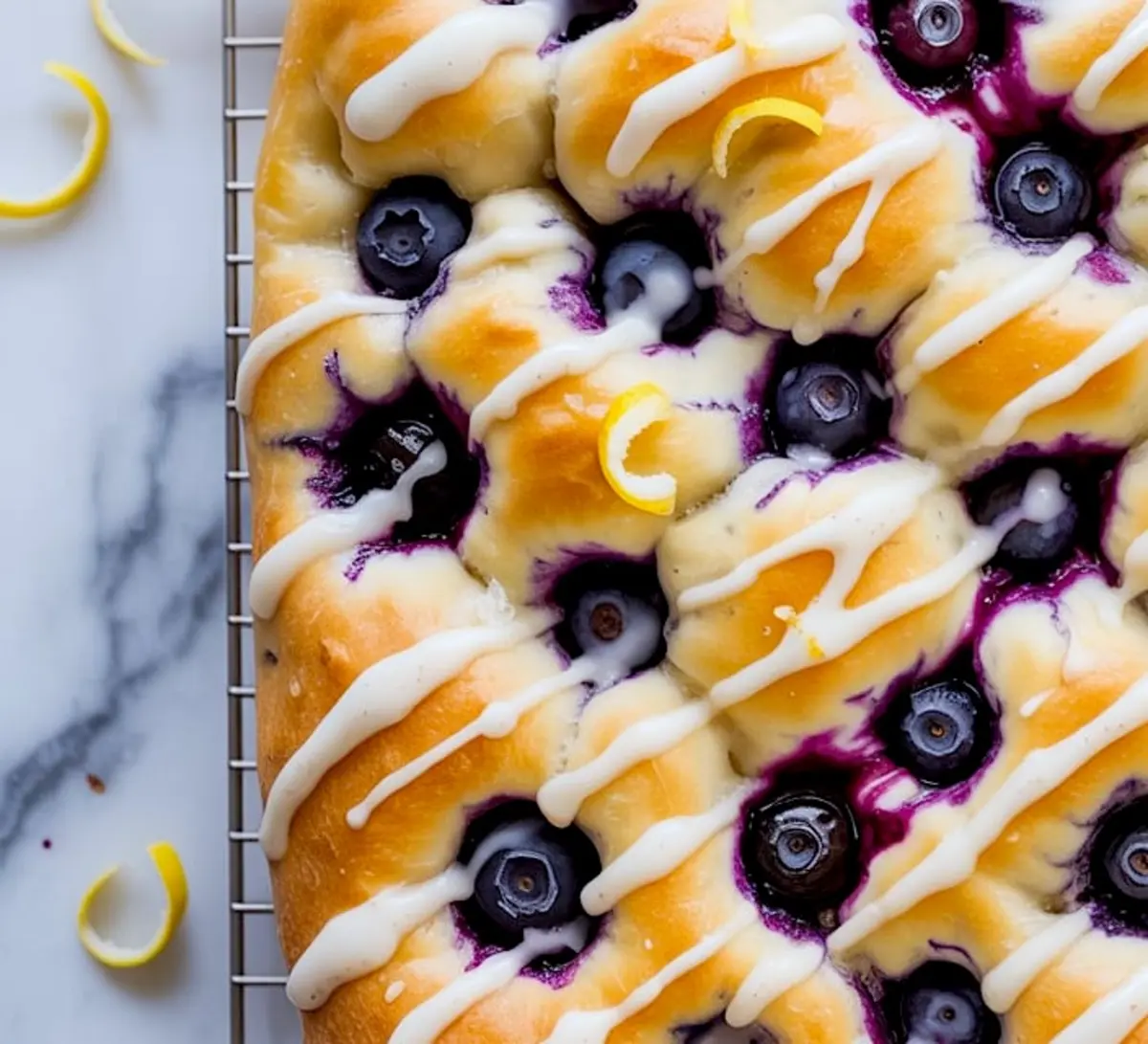 Golden baked lemon blueberry focaccia on a wire rack, topped with fresh blueberries, icing drizzle, and thin lemon peel curls over a marble surface.
