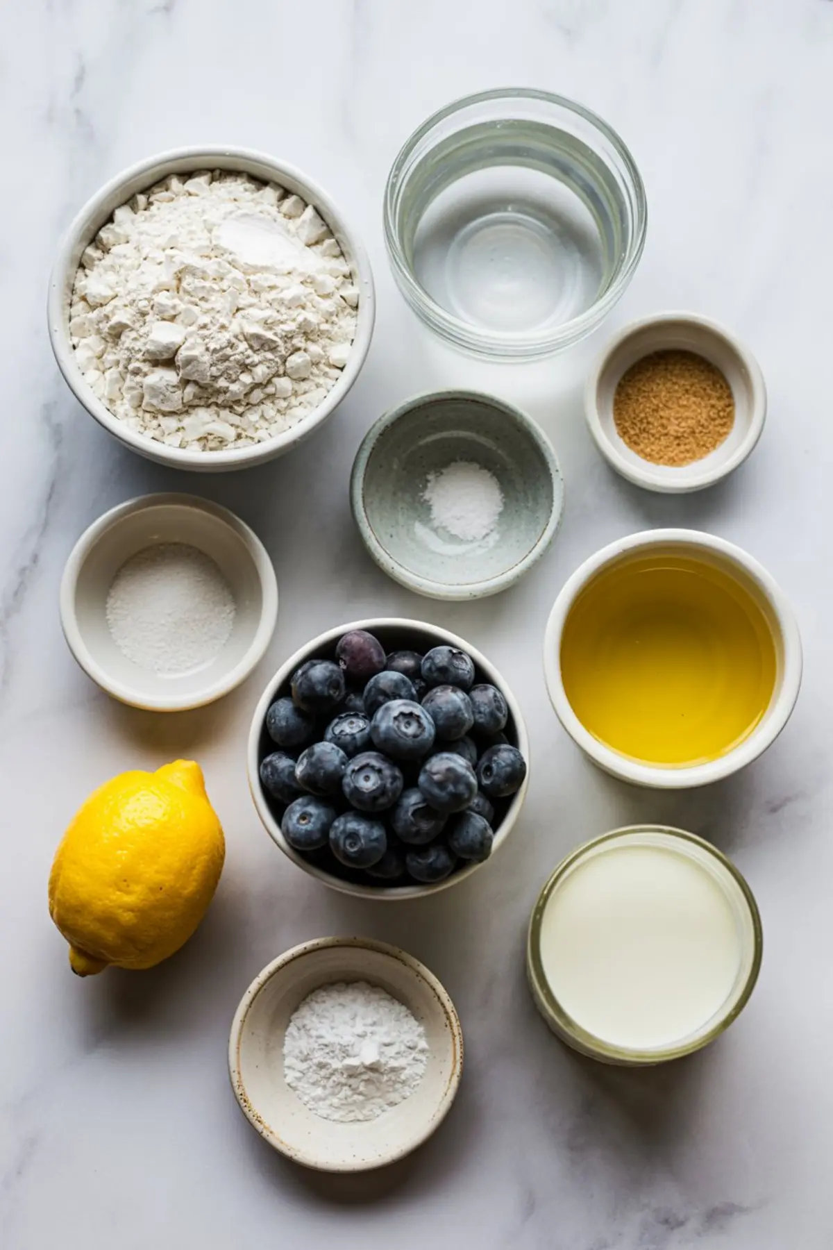 Flat lay of ingredients for lemon blueberry focaccia including all-purpose flour, fresh blueberries, lemon, olive oil, powdered sugar, yeast, and milk.
