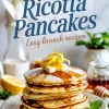 Stack of lemon ricotta pancakes drizzled with syrup and topped with butter and lemon zest, with a honey dipper and sliced lemons in the background on a marble surface.