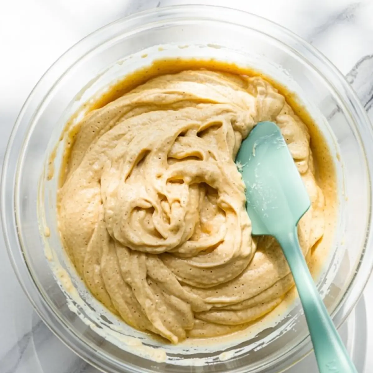 Smooth and airy lemon ricotta pancake batter in a clear bowl with a teal silicone spatula on a light marble countertop.
