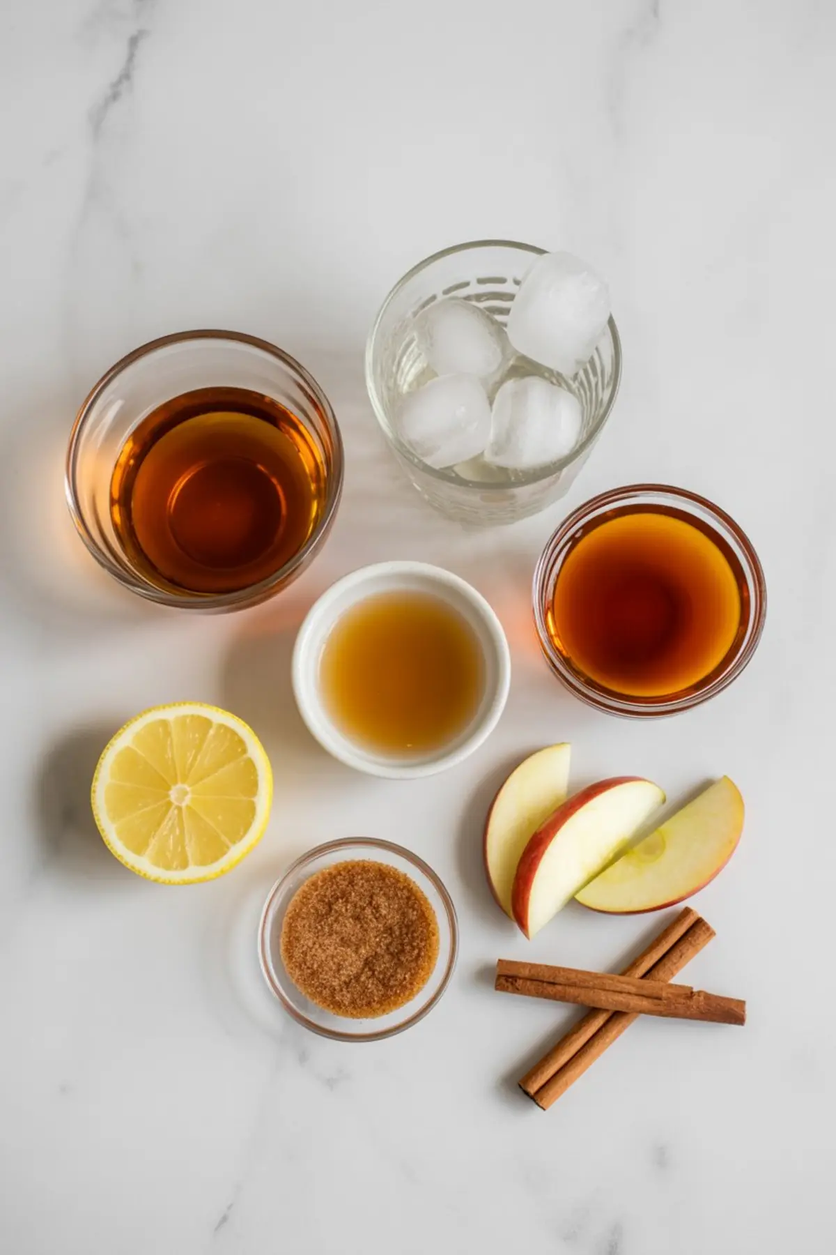 Flat lay of ingredients for a maple bourbon apple cider cocktail, including lemon halves, apple slices, brown sugar, cinnamon sticks, maple syrup, apple cider, bourbon, and a glass of ice cubes on a marble surface.
