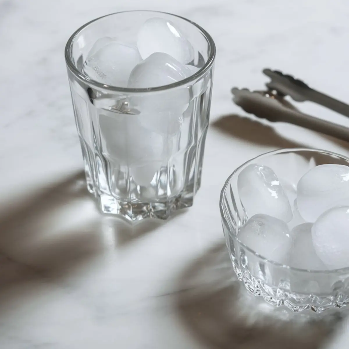 Close-up of a glass and bowl filled with ice cubes on a marble surface, accompanied by metal ice tongs for drink preparation.
