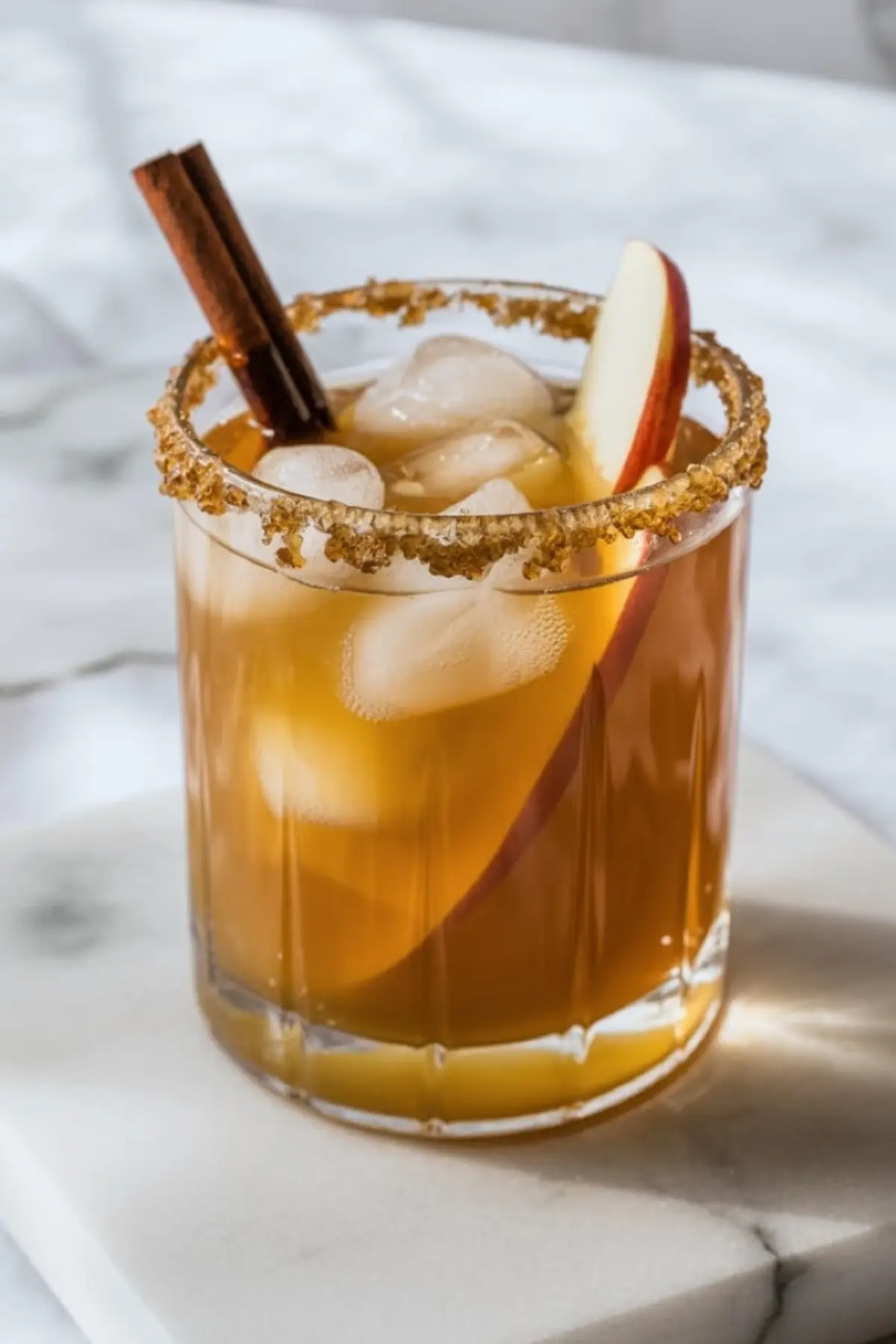 Glass of maple bourbon apple cider cocktail with ice, garnished with a cinnamon stick and apple slice, and rimmed with brown sugar on a marble coaster.
