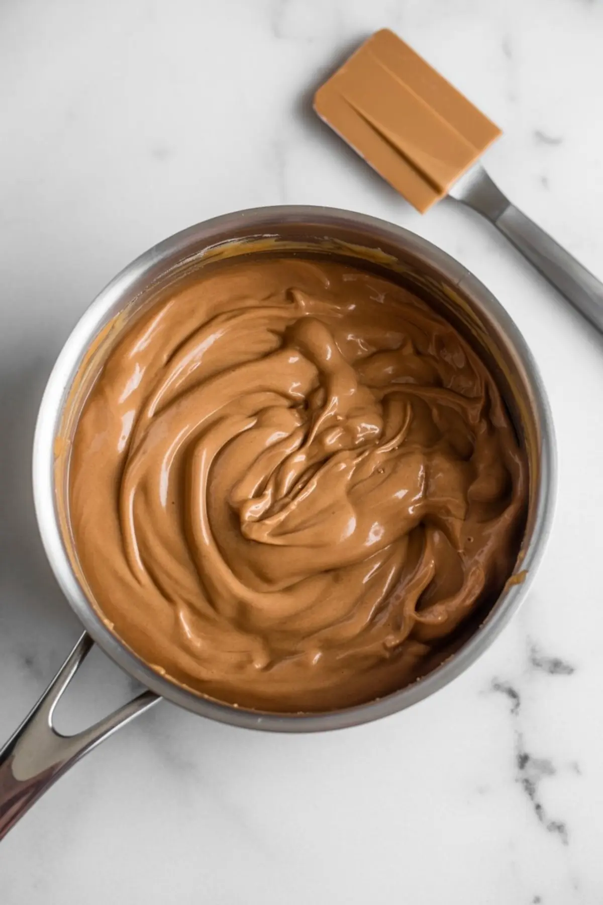 Saucepan filled with smooth, creamy maple fudge mixture, freshly stirred with a spatula placed beside it on a marble surface.