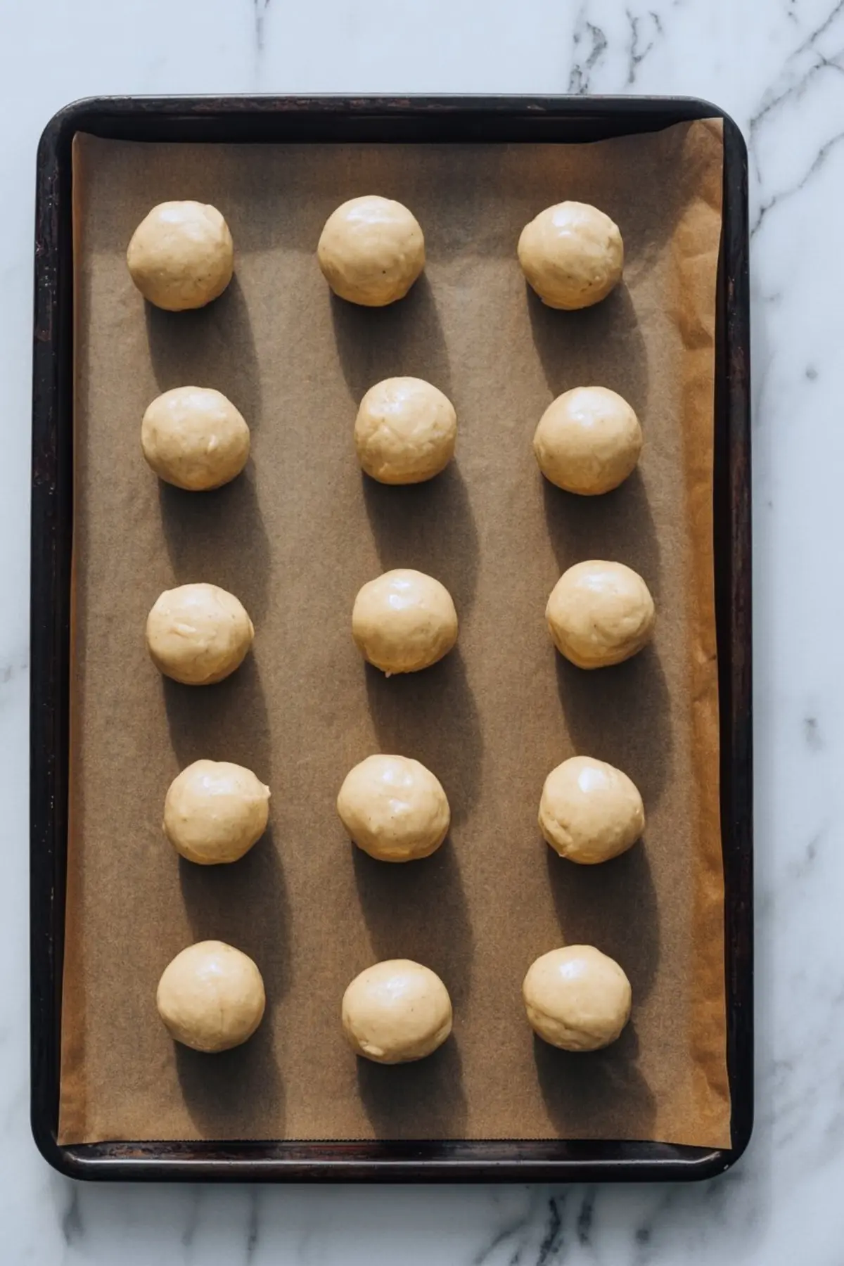 Brown parchment-lined sheet pan shows neat rows of portioned sugar cookie dough balls ready to bake into melting snowman cookie bases.