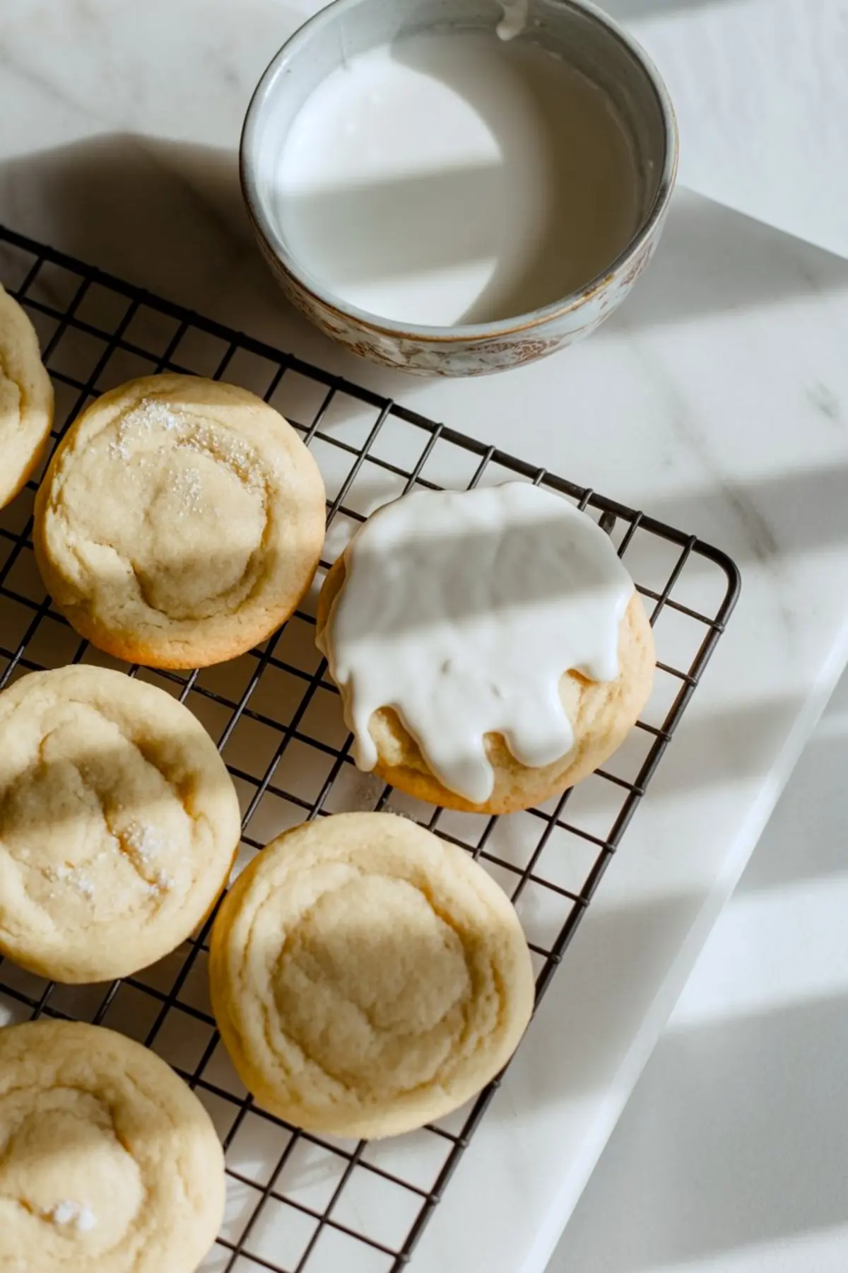 Wire rack holds freshly baked sugar cookie bases for melting snowman cookies, one dripping with glossy vanilla icing beside a ceramic bowl of glaze on a bright marble countertop.