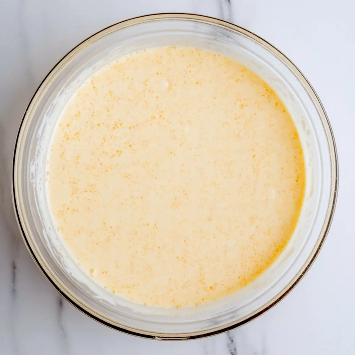 Top-down view of a mixing bowl containing light yellow tres leches milk mixture made with evaporated milk, condensed milk, and cream, used to soak sponge cake layers.
