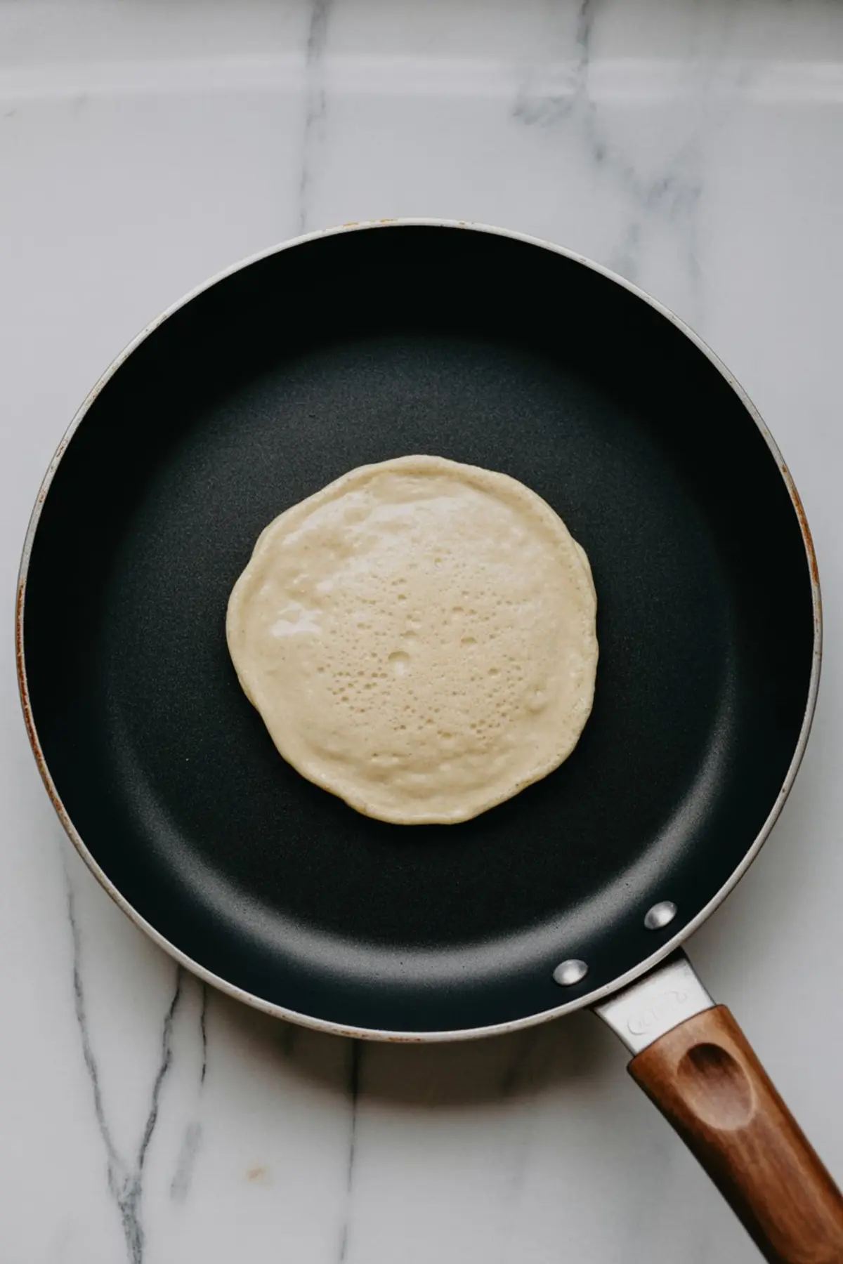 Oat pancake cooking in a non-stick skillet with a wooden handle on a marble countertop, showing bubbles forming on the surface.