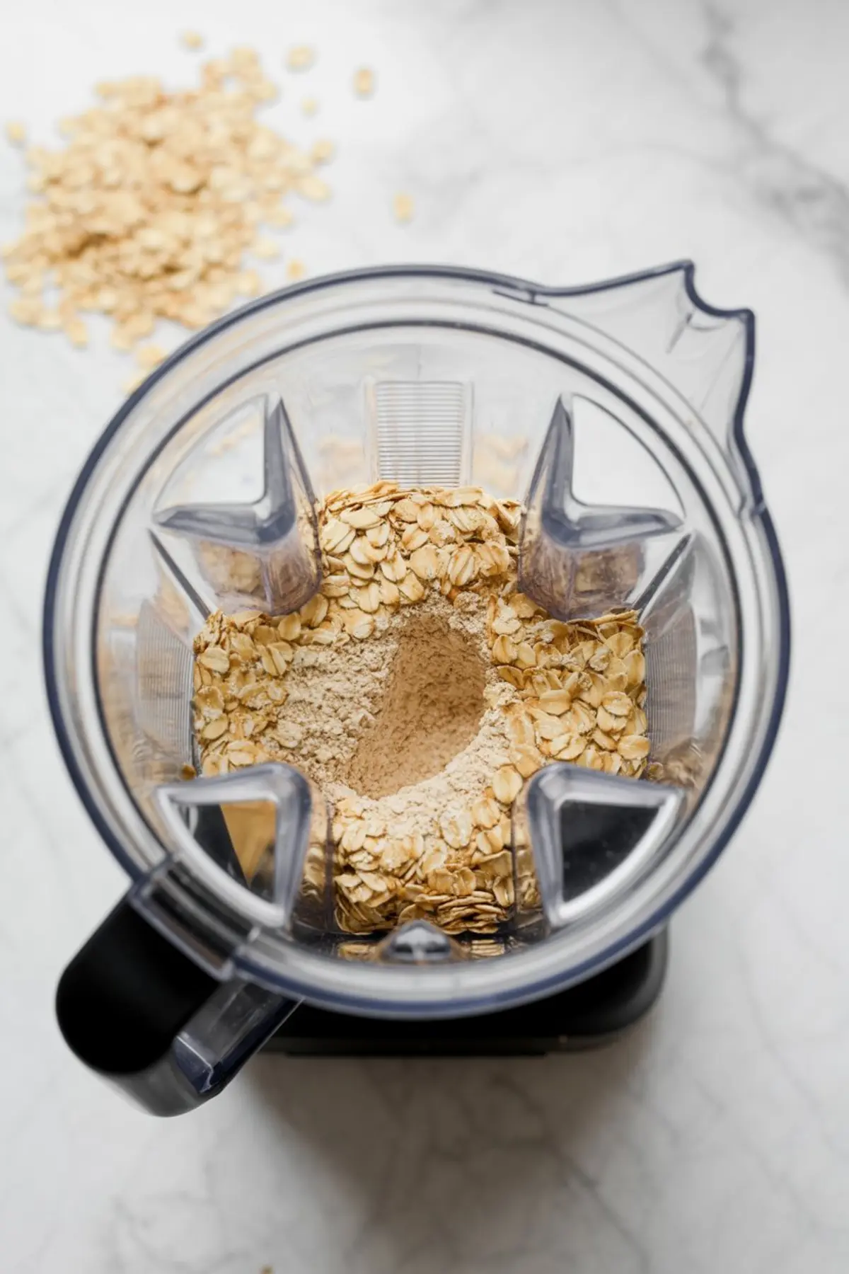 Top-down view of rolled oats and oat flour inside a blender on a marble surface, with loose oats scattered in the background.