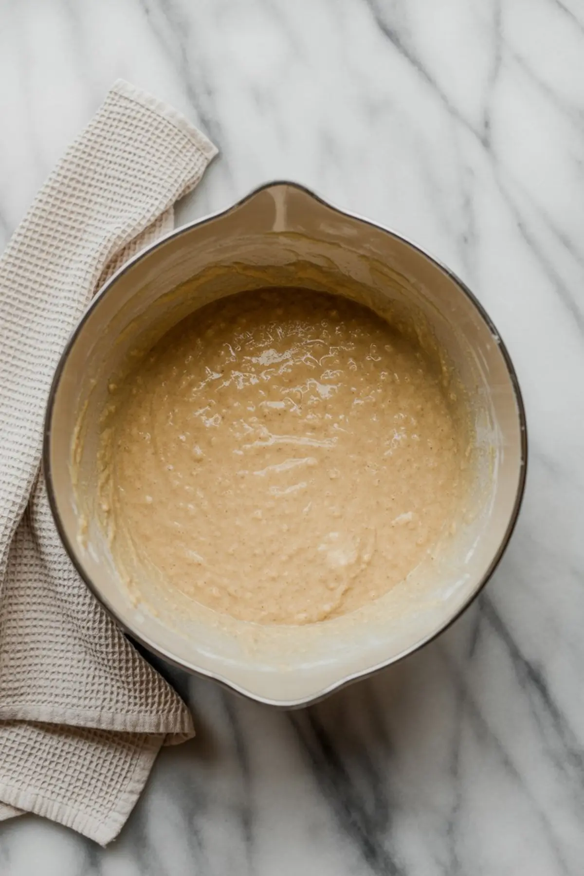 Creamy oat pancake batter in a mixing bowl placed on a marble surface with a folded beige kitchen towel beside it.