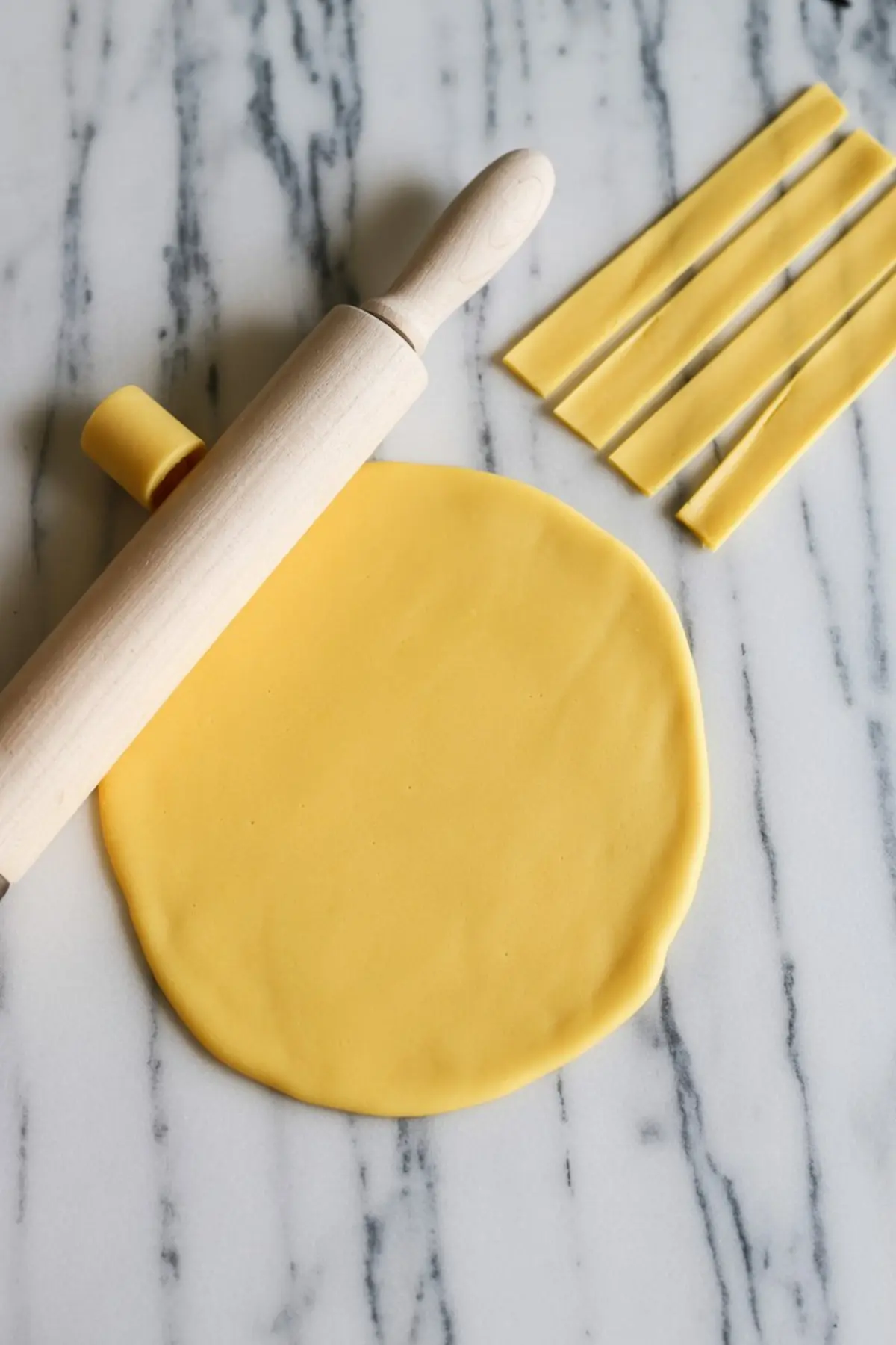 Rolled out yellow fondant on a marble surface with several uniform strips cut, alongside a wooden rolling pin.