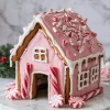 Assembled pink gingerbread house with white icing details, peppermint sticks for columns, and crushed peppermint candy topping, placed on a round marble board with holiday greenery.