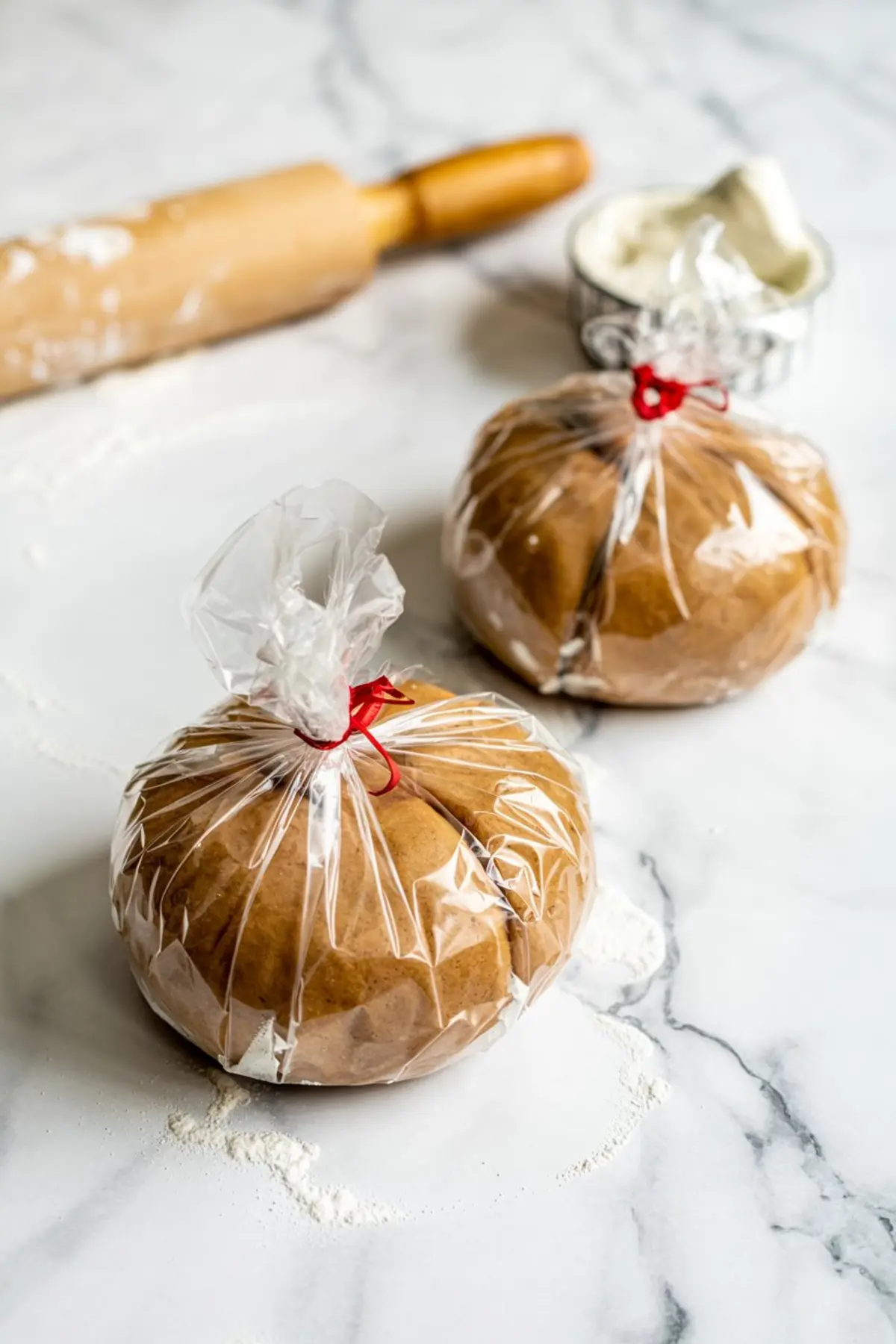 Two portions of gingerbread dough wrapped in plastic and tied with red ribbons, placed on a lightly floured marble surface near a rolling pin and bowl of flour.