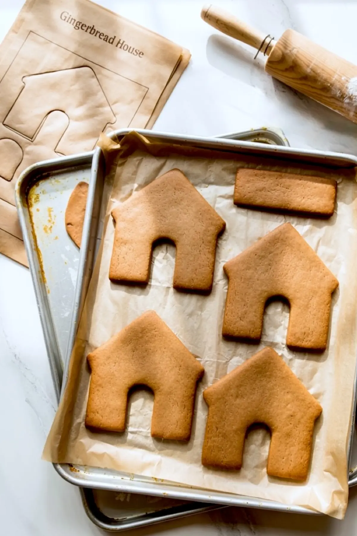 Baking sheet with freshly baked gingerbread house pieces laid on parchment paper, including front and side panels, with a rolling pin, ruler, and house template on a marble countertop.