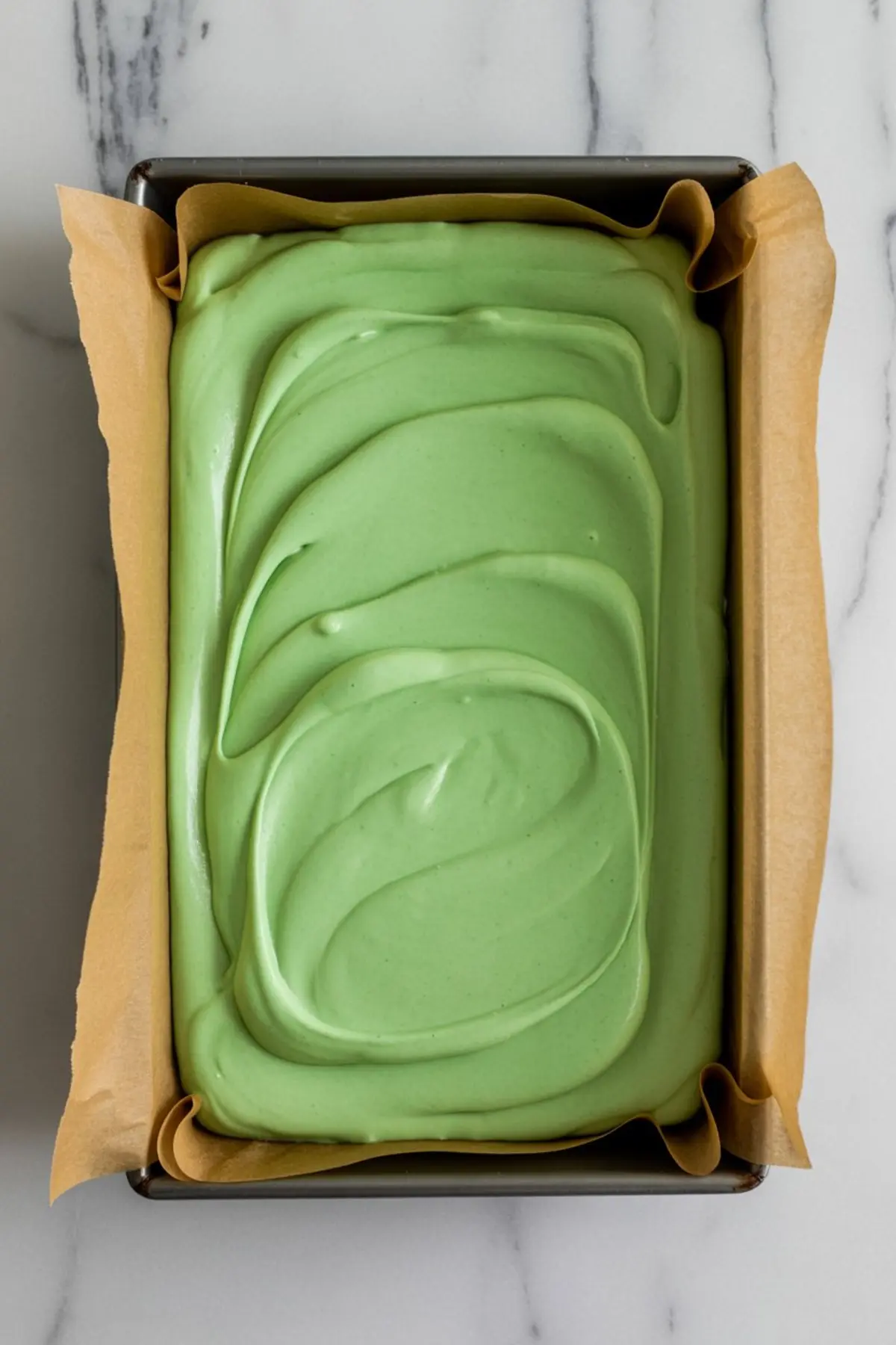 Green fudge mixture spread in a parchment-lined rectangular pan, with smooth swirls on top. Step in making layered Halloween fudge before chocolate drizzle is added.
