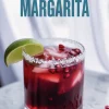 Bright red pomegranate margarita in a short glass with coarse salt rim, ice cubes, and floating pomegranate seeds, garnished with a fresh lime wedge and set against a soft curtain backdrop.