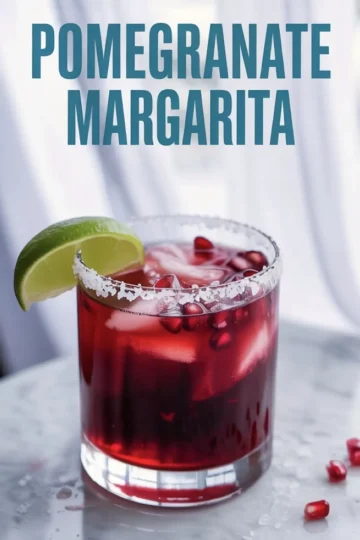Bright red pomegranate margarita in a short glass with coarse salt rim, ice cubes, and floating pomegranate seeds, garnished with a fresh lime wedge and set against a soft curtain backdrop.