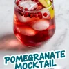 Single glass of red pomegranate mocktail garnished with lime wedge and pomegranate seeds, placed on a white marble surface with a vibrant mocktail label overlay at the bottom.