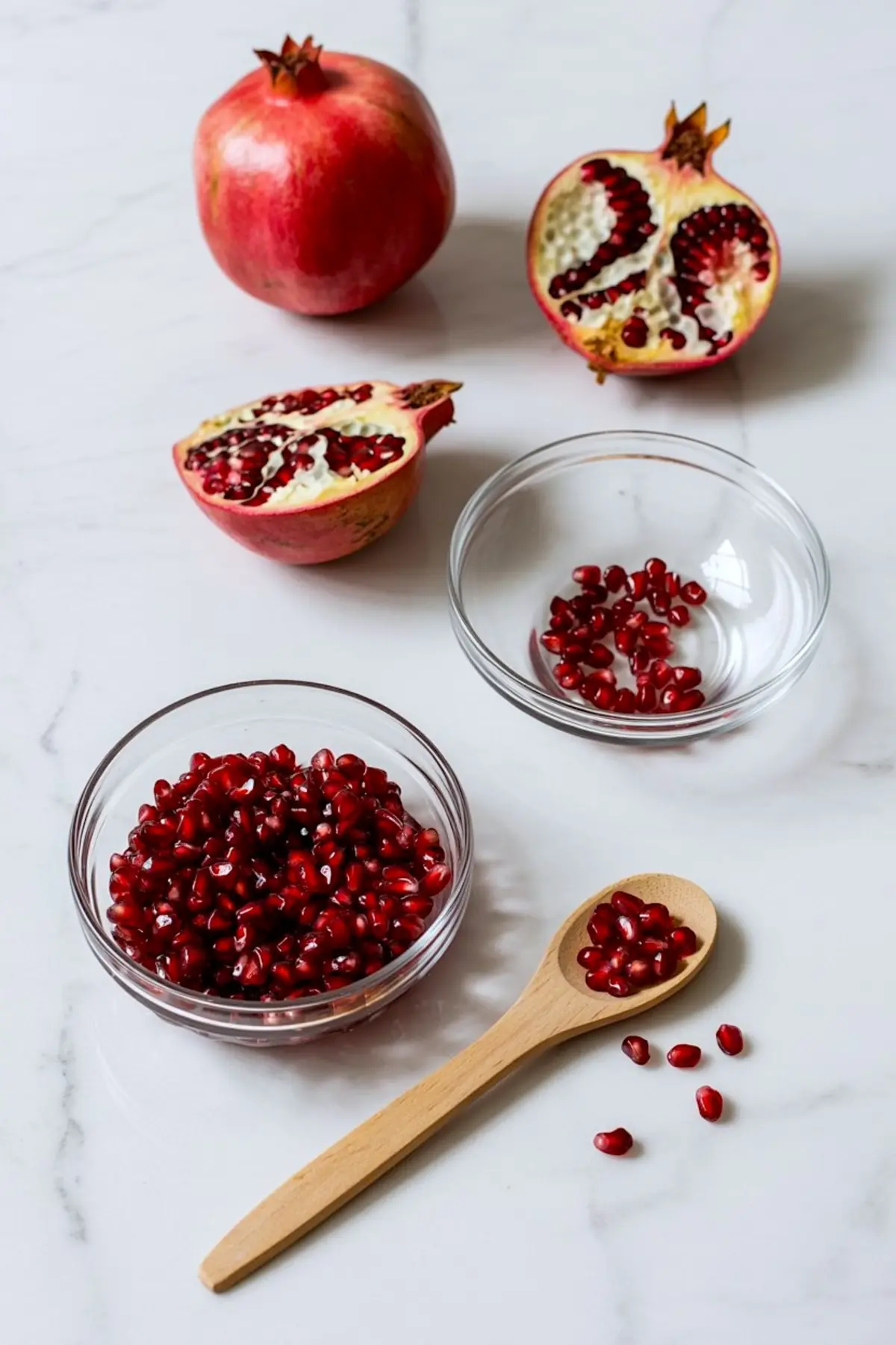Arrangement of whole and halved pomegranates, along with bowls and a wooden spoon filled with fresh pomegranate arils, styled on a white marble surface for an ingredient prep display.